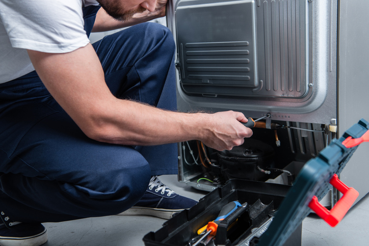 partial view of repairman in working overall fixing refrigerator by screwdriver in kitchen