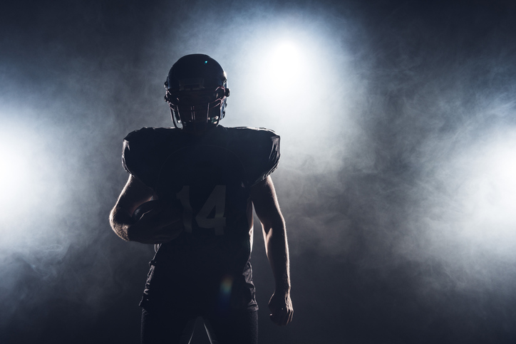 dark silhouette of equipped american football player with ball against white smoke