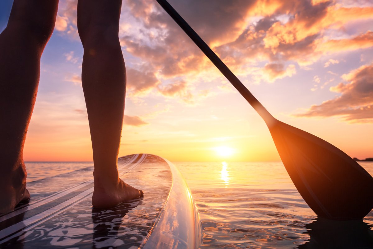 Stand up paddle boarding or standup paddleboarding on quiet sea at sunset with beautiful colors during warm summer beach vacation holiday, active woman, close-up of water surface, legs and board