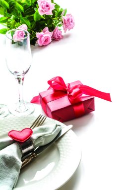 Valentines day dinner with table place setting with red gift, glass for champagne, heart ornaments with silverware on white. Close up. Valentine’s card.