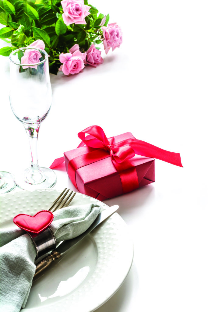 Valentines day dinner with table place setting with red gift, glass for champagne, heart ornaments with silverware on white. Close up. Valentine’s card.