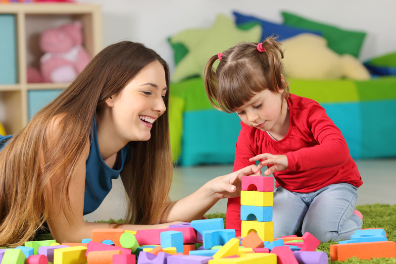 Mother and daughter playing together with colorful construction toys on a carpet on the floor af a bedroom at home teen babysitting