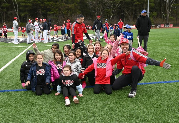Young Little League players throw their hands in the air while sitting on the ground, smiling with their coaches on a field.