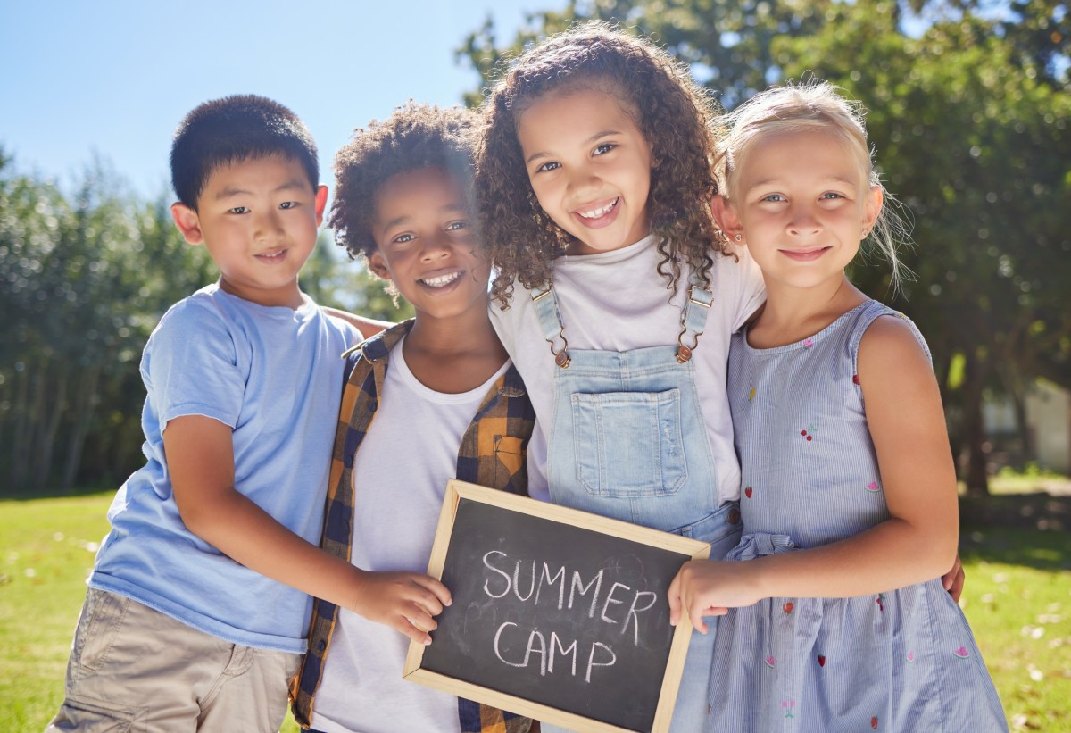 A group of summer camp friends holding a sign in a outside in a park