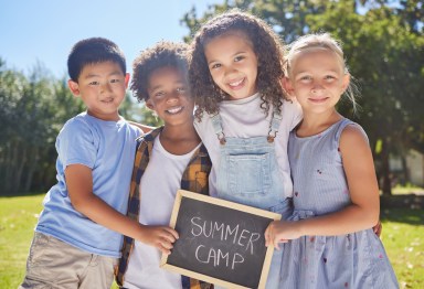 A group of summer camp friends holding a sign in a outside in a park