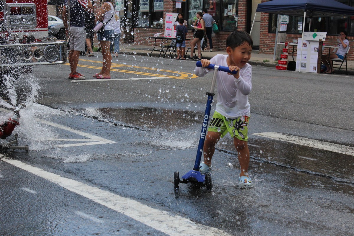A child rides his scooter through a sprinkler during the Al Fresco on Plandome Road.