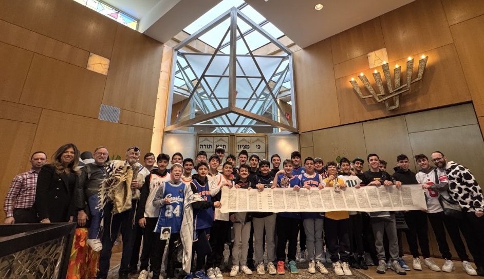 (L-R) Cantor Yitzy Spinner; Laura Cohen, NSHA Exec. Director; Dr. Jeffrey Kobrin, NSHA Rosh HaYeshiva/Head of School; Dr. Paul Brody, Megillah Program initiator, holding Megillah & his Grandfather’s Prayer Shawl; NSHA Middle School 2026 Megillah Readers, in Purim costumes, proudly unfurling the “Dr. Paul Brody Megillah;” at far right, Rabbi Simon Basalely, Middle School Judaic Studies Principal and Sephardic Megillah instructor. A composite of the latest “Contemporary “Hamans,” from the Gaza & Iran Wars,” created by Drora Brody, swung when mentioning Haman & his 10 sons hanging from the tree - is held by Jack Muller (6th from left), son of Daniella Muller, president of NSHA, and others, held over the Megillah.